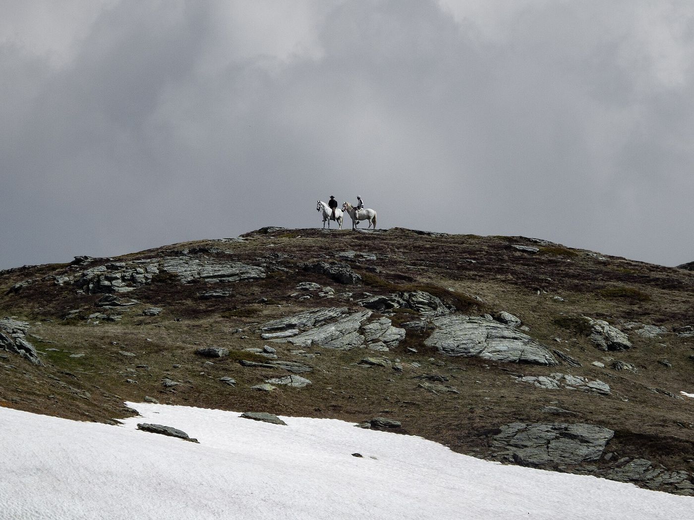 Two riders on a high rocky ridge with snow patches